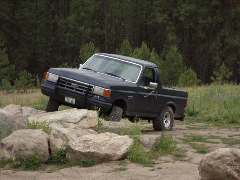 1988 Ford Bronco - A dark blue Ford Bronco is shown driving over large rocks on a dirt path, with a forest in the background. The vehicle is angled to the rig…