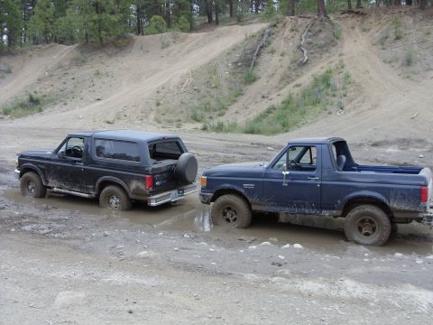 1988 Ford Bronco - Two mud-splashed full-size Ford Broncos parked nose-to-tail in a puddle on a dirt trail: a black one with its hardtop and rear-mounted spar…