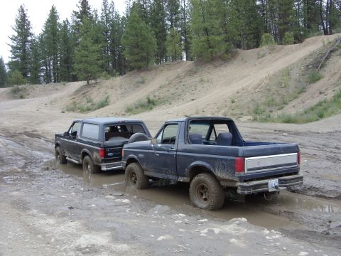 1988 Ford Bronco - Two mud-splattered full-size Ford Broncos parked nose-to-tail in a wet, rutted dirt pit surrounded by pine trees. The nearer dark blue Bron…