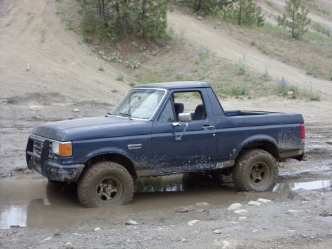 1988 Ford Bronco - A dark blue Ford Bronco is stuck in a muddy puddle on a dirt road with trees and a sandy hill in the background.