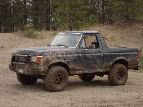 1988 Ford Bronco - A dark blue, mud-splattered Ford Bronco is parked on a dirt road with a wooded hillside in the background. The vehicle has large off-road t…