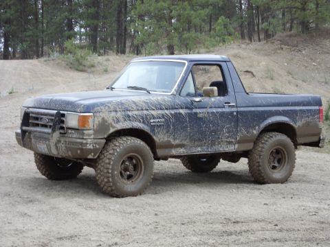 1988 Ford Bronco - Driver-side 3/4 front view of a dark blue full-size Ford Bronco with the rear hardtop removed, parked on a dirt pad in a pine forest. Mud i…