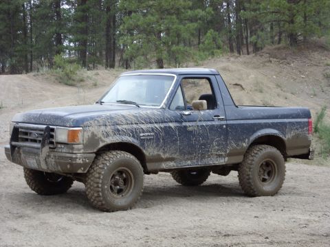 1988 Ford Bronco - Driver-side view of a dark blue full-size Ford Bronco caked in dried mud splatter, top removed to leave the windshield frame and roll bar e…