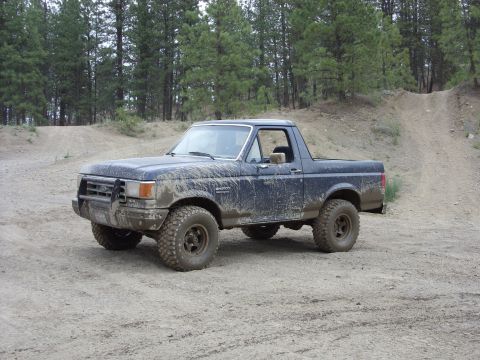 1988 Ford Bronco - A dark blue, mud-covered Ford Bronco is parked on a dirt road with a forest in the background. The vehicle has a front brush guard and off-…