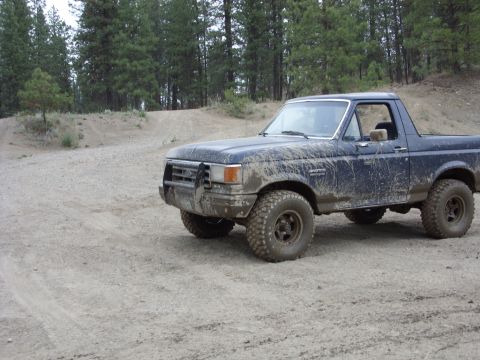1988 Ford Bronco - A muddy, dark blue Ford Bronco is parked on a dirt road with trees in the background. The vehicle has large off-road tires and appears to b…