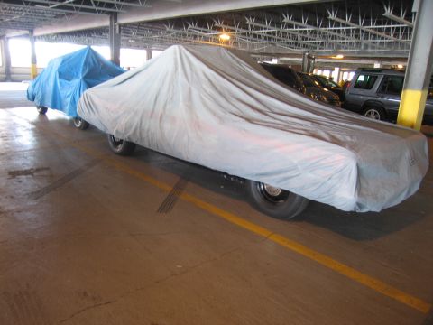 1991 Ford LTD Crown Victoria - sedan draped in a gray car cover sits beside a blue-tarped vehicle on a wet garage floor.