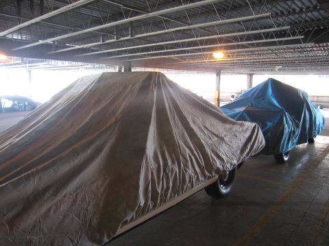1991 Ford LTD Crown Victoria - Two large vehicles draped in brown and teal car covers parked in a covered parking structure.