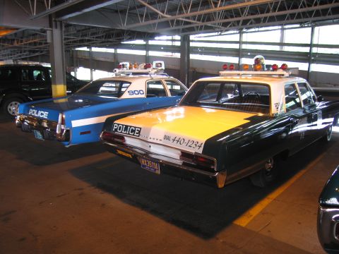 1991 Ford LTD Crown Victoria - Rear view of a green-and-yellow POLICE sedan lettered DIAL 440-1234 beside a blue-and-white patrol car numbered 900 in a garage.