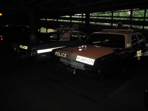 1991 Ford LTD Crown Victoria - dim row of vintage POLICE patrol cars in a garage, including a tan-and-black sedan lettered DIAL 440-1234.
