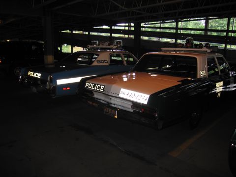 1991 Ford LTD Crown Victoria - tan-and-black POLICE sedan lettered DIAL 440-1234 parked beside a blue-and-white patrol car numbered 900 in a dim garage.