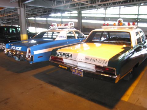 1991 Ford LTD Crown Victoria - Rear view of a yellow-and-green POLICE sedan lettered DIAL 440-1234 beside a blue-and-white patrol car numbered 900 indoors.