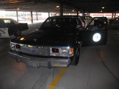 1991 Ford LTD Crown Victoria - black Ford full-size sedan with a front door open revealing a glowing white circular emblem, parked in a garage.