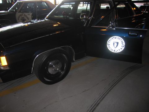 1991 Ford LTD Crown Victoria - black sedan with its door open, displaying a brightly illuminated circular police seal emblem, on a tire-marked garage floor.