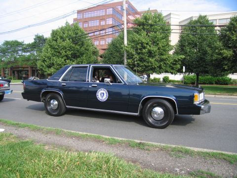1991 Ford LTD Crown Victoria - dark blue full-size sedan bearing a circular state seal emblem on the door drives along a tree-lined street past office buildings.