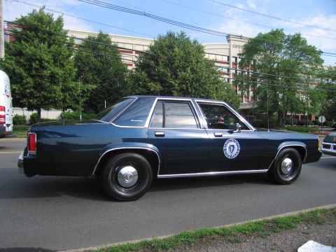 1991 Ford LTD Crown Victoria - Side profile of a dark blue full-size four-door sedan with a circular seal on the door, parked along a roadway by trees.