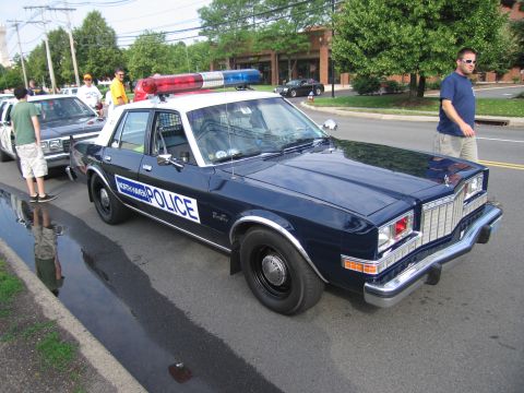 1991 Ford LTD Crown Victoria - dark blue POLICE sedan with a red roof light bar parked on a street as bystanders stand near it under power lines.