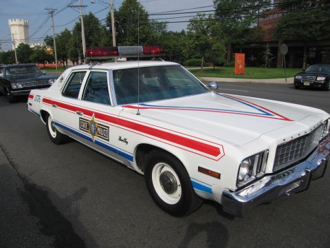 1991 Ford LTD Crown Victoria - white Plymouth four-door marked STATE POLICE with red and blue stripes and a roof light bar parked outdoors near trees.