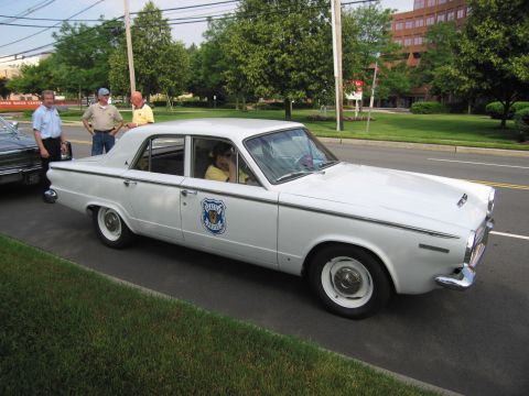 1991 Ford LTD Crown Victoria - white early-1960s four-door sedan wearing a blue-and-white police door shield, parked roadside with three men standing nearby.