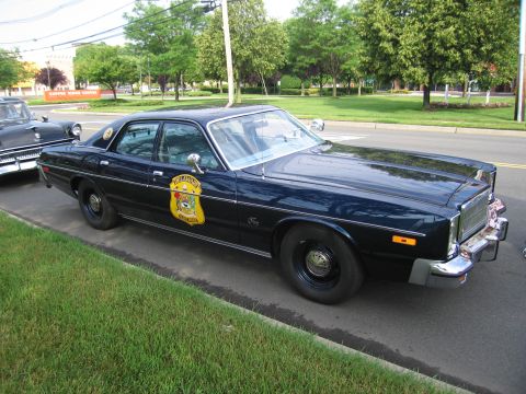 1991 Ford LTD Crown Victoria - black mid-1970s four-door police sedan with a gold state-police door shield and roof beacon, parked beside a grass curb.