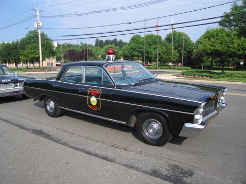 1991 Ford LTD Crown Victoria - black early-1960s full-size four-door police sedan with a gold-and-black door shield and red roof beacon, parked on pavement.