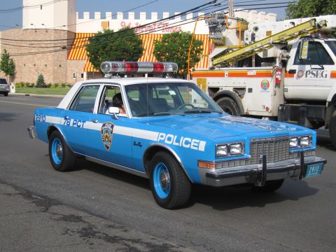1991 Ford LTD Crown Victoria - light blue 1980s four-door police sedan in NYPD livery lettered POLICE and 78 PCT with red roof lightbar, parked near a utility truck.
