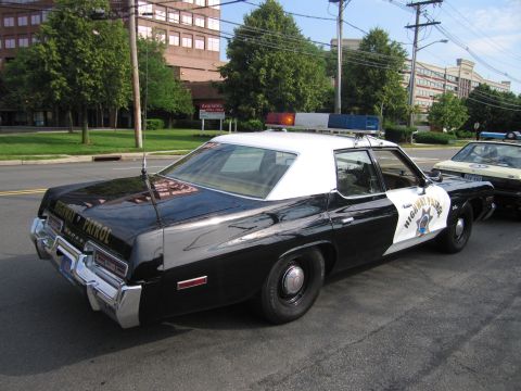 1991 Ford LTD Crown Victoria - Rear three-quarter view of a black 1970s highway patrol sedan with white roof, door star shield and roof beacon, parked on a street by buil…