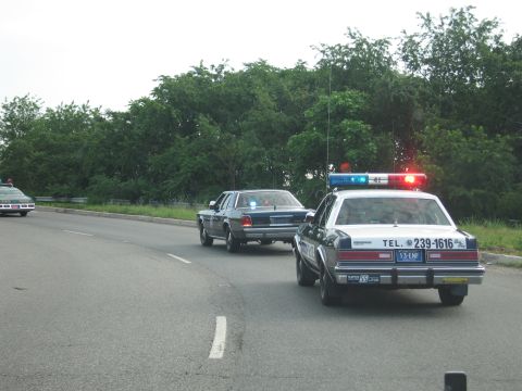 1991 Ford LTD Crown Victoria - Two police cars driving on a curving road; the trailing white cruiser is lettered TEL. 239-1616 with lit red-and-blue roof lights.