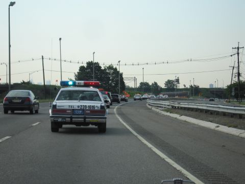 1991 Ford LTD Crown Victoria - white police cruiser lettered TEL. 239-1616 with a lit blue roof light driving on a multi-lane highway, a dark car passing on the left.