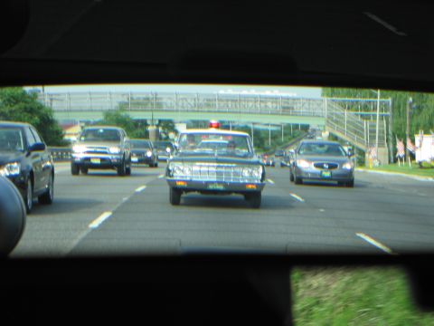 1991 Ford LTD Crown Victoria - Windshield-framed view of a dark vintage sedan with a red roof beacon driving on a highway beneath an overpass, traffic in adjacent lanes.