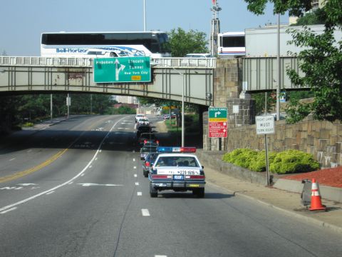 1991 Ford LTD Crown Victoria - white police cruiser with roof lights driving on a road toward an overpass marked with Hoboken and Lincoln Tunnel highway signs.