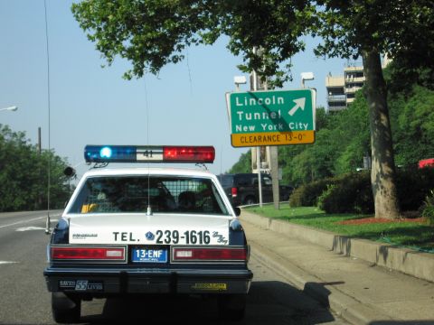 1991 Ford LTD Crown Victoria - Rear view of a white police cruiser lettered TEL. 239-1616 with lit roof lights, stopped beside a green Lincoln Tunnel, New York City sign.