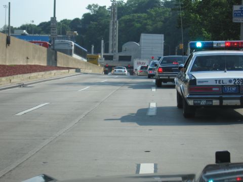 1991 Ford LTD Crown Victoria - white police cruiser lettered TEL. 239 with lit roof lights driving on a wide divided roadway toward a tunnel entrance, trucks ahead.