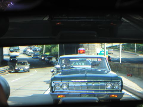 1991 Ford LTD Crown Victoria - Windshield-framed view of a dark vintage sedan with a red roof beacon leading a line of cars on a road toward a stone overpass.