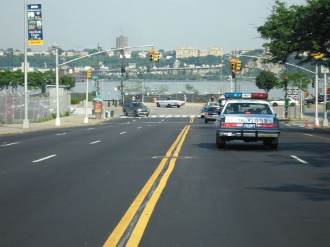 1991 Ford LTD Crown Victoria - police-style sedan with a roof light bar drives down a wide multi-lane road heading toward a river and distant bridge.