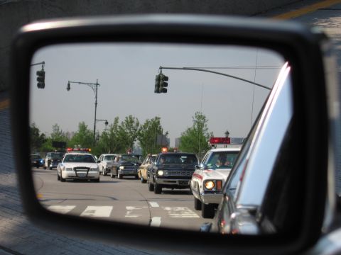 1991 Ford LTD Crown Victoria - car's side mirror reflects a queue of vehicles, including police-style cars with light bars, waiting at a city traffic light.