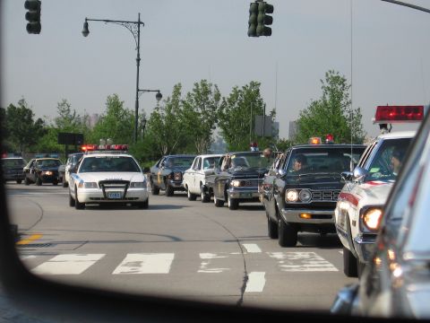 1991 Ford LTD Crown Victoria - convoy of older sedans and police-style cars with light bars is stopped in traffic at a tree-lined city intersection.