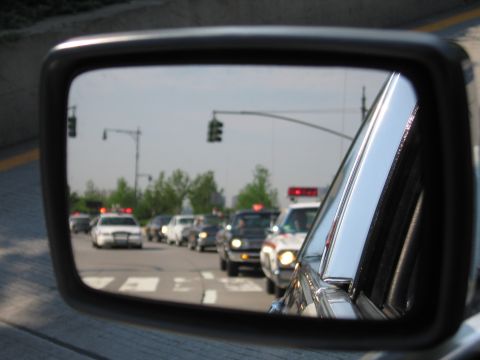 1991 Ford LTD Crown Victoria - side mirror reflects a row of cars and police-style sedans with red lights driving on a city street near a crosswalk.