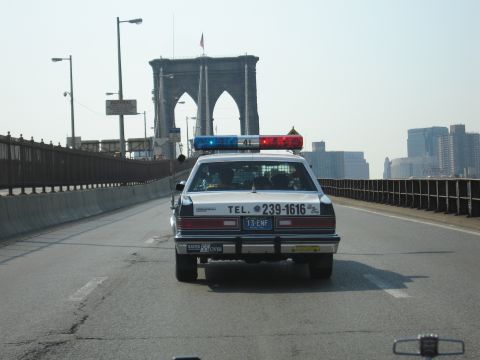1991 Ford LTD Crown Victoria - rear of a black-and-white police-style sedan with a light bar and TEL. 239-1616 lettering drives across the Brooklyn Bridge.