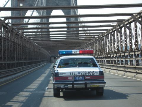 1991 Ford LTD Crown Victoria - black-and-white police-style sedan marked TEL. 239-1616 drives through the steel trusswork of a bridge roadway.