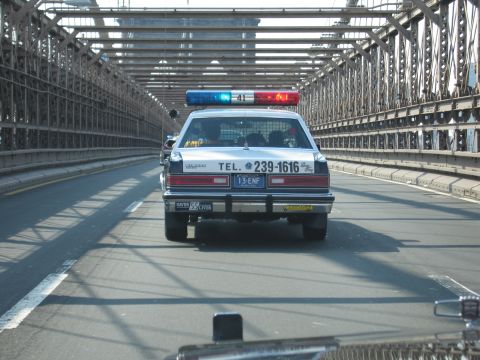 1991 Ford LTD Crown Victoria - rear of a black-and-white police-style sedan with a lit roof bar moves along a road enclosed by steel bridge trusses.