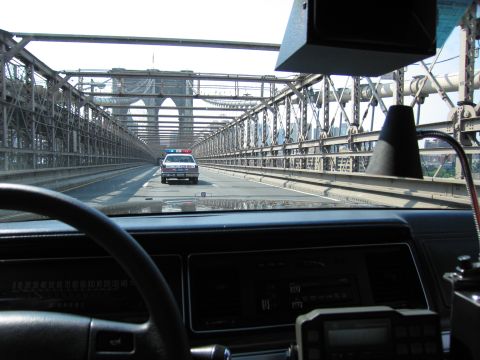 1991 Ford LTD Crown Victoria - Seen over a car's dashboard and steering wheel, a police-style sedan drives ahead through a steel-truss bridge roadway.