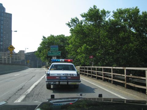 1991 Ford LTD Crown Victoria - black-and-white police-style sedan with a light bar drives along an elevated city highway lined with trees and a wood guardrail.