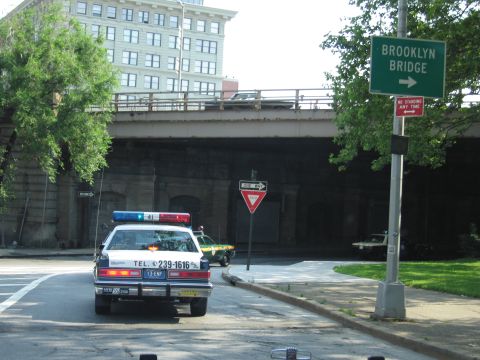 1991 Ford LTD Crown Victoria - police-style sedan waits at an intersection under a stone overpass beside a green Brooklyn Bridge directional sign.