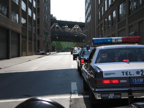 1991 Ford LTD Crown Victoria - black-and-white police-style sedan with Connecticut plates leads a line of cars down a narrow street between tall buildings under an overpa…
