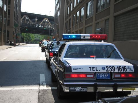1991 Ford LTD Crown Victoria - black-and-white Plymouth police-style sedan marked TEL. 239-1616 with Connecticut plates sits in a line of cars on an urban street.
