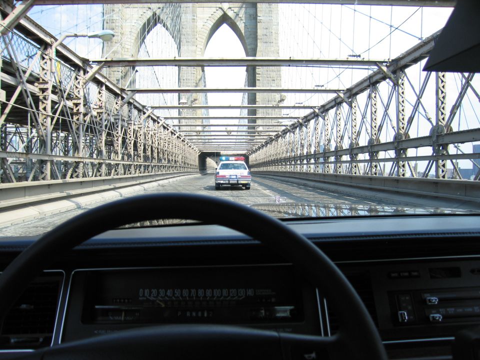 Windshield view of a police-style car driving ahead through Brooklyn Bridge cables and trusses