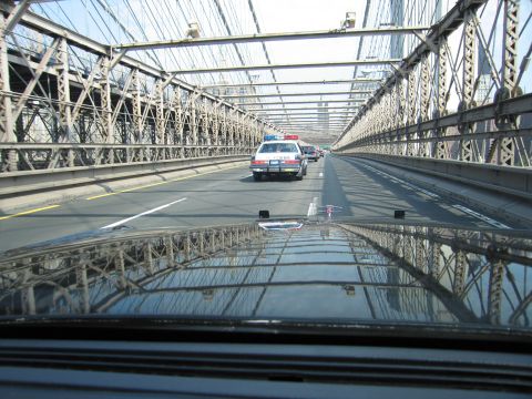 1991 Ford LTD Crown Victoria - police-style sedan drives ahead on a steel suspension bridge, its surroundings mirrored in the dark hood of the trailing car.