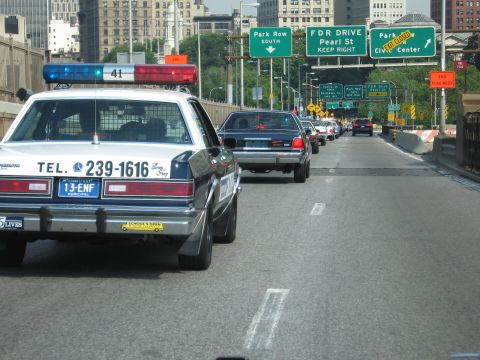 1991 Ford LTD Crown Victoria - black-and-white police-style sedan marked TEL. 239-1616 leads traffic on a city expressway beneath green FDR Drive and Pearl St signs.