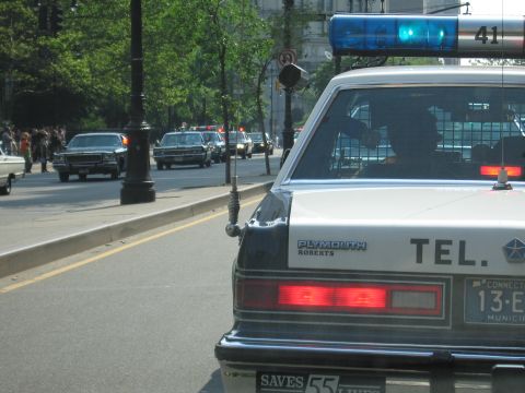 1991 Ford LTD Crown Victoria - rear of a black-and-white Plymouth police-style sedan with a lit roof bar and Connecticut plate, parked on a sunny tree-lined city street.