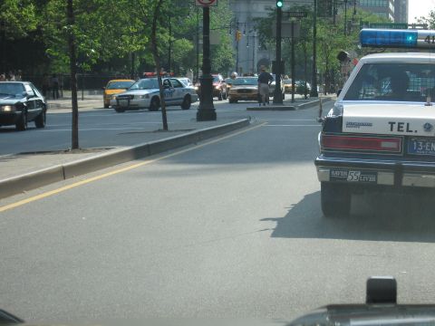 1991 Ford LTD Crown Victoria - black-and-white police-style sedan drives a city street as an NYPD Crown Victoria and a yellow taxi sit across the tree-lined median.